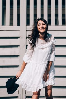 Sunny lifestyle fashion portrait of young stylish hipster woman walking on the street, wearing trendy white dress, black hat and boots. Gray wooden backgrond.