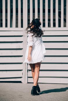 Sunny lifestyle fashion portrait of young stylish hipster woman walking on the street, wearing trendy white dress, black hat and boots. Gray wooden backgrond.