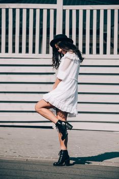 Sunny lifestyle fashion portrait of young stylish hipster woman walking on the street, wearing trendy white dress, black hat and boots. Gray wooden backgrond.