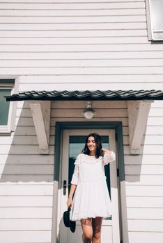 Sunny lifestyle fashion portrait of young stylish hipster woman walking on the street, wearing trendy white dress, black hat and boots. White wooden house on a backgrond.