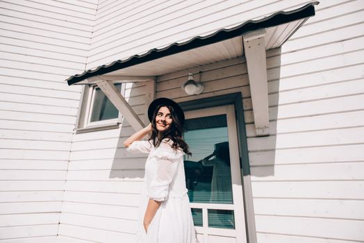 Sunny lifestyle fashion portrait of young stylish hipster woman walking on the street, wearing trendy white dress, black hat and boots. White wooden house on a backgrond.