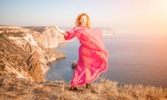 Side view a Young beautiful sensual woman in a red long dress posing on a volcanic rock high above the sea during sunset. Girl on the nature on blue sky background. Fashion photo