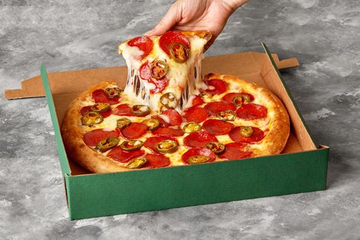 Female hand taking slice of appetizing fresh pizza with melted mozzarella cheese, pepperoni sausage and spicy jalapeno peppers lying in carton box on gray stone surface. Popular fast food concept