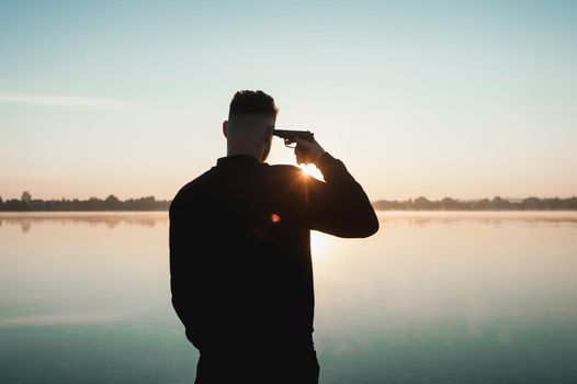 Suicide minds. silhouette of depressed young man holding gun near his head standing near water.