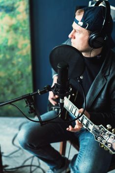 Close up of a man singer in a headphones with a guitar recording a track in a home studio. Man wearing jeans black shirt and a jacket. side view