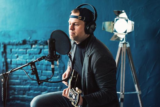 Close up of a man singer in a headphones with a guitar recording a track in a home studio. Man wearing jeans black shirt and a jacket.