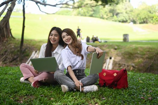 Two cheerful asian women sitting together in park on the green grass.