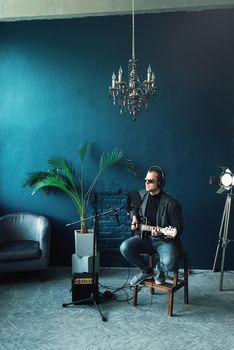 Close up of a man singer in a headphones with a guitar recording a track in a home studio. Man wearing sunglasses, jeans, black shirt and a jacket.