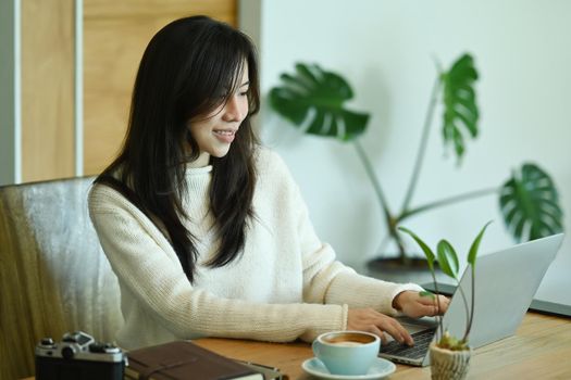 Thoughtful asian woman in white sweater using laptop computer at home.