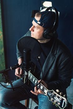 Close up of a man singer in a headphones with a guitar recording a track in a home studio. Man wearing jeans black shirt and a jacket. side view