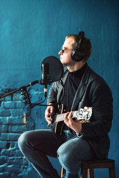 Close up of a man singer in a headphones with a guitar recording a track in a home studio. Man wearing sunglasses, jeans, black shirt and a jacket. side view