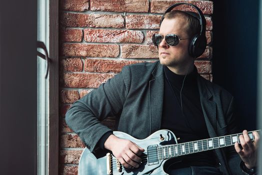 Man singer sitting on a window sill in a headphones with a guitar recording a track in a home studio. Man wearing sunglasses, jeans, black shirt and a jacket.