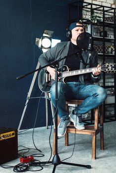 Close up of a man singer in a headphones with a guitar recording a track in a home studio. Man wearing jeans black shirt and a jacket.