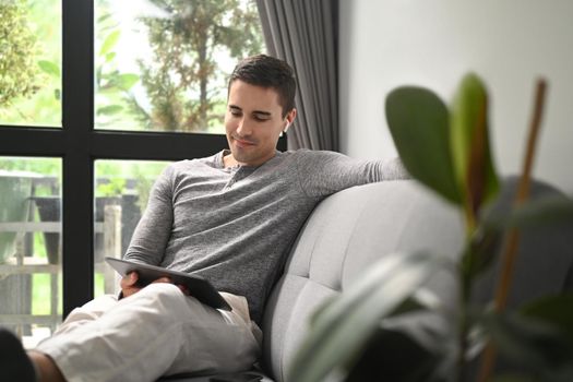 Happy man using digital tablet while sitting on comfortable sofa.