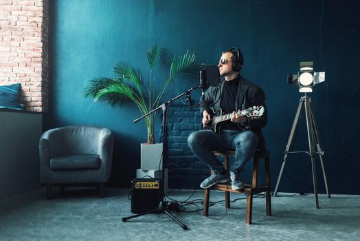 Close up of a man singer in a headphones with a guitar recording a track in a home studio. Man wearing sunglasses, jeans, black shirt and a jacket.