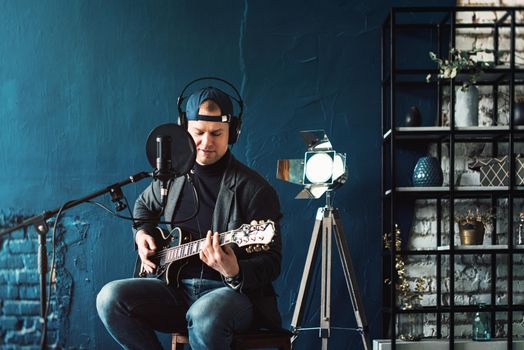 Close up of a man singer in a headphones with a guitar recording a track in a home studio. Man wearing jeans black shirt and a jacket.