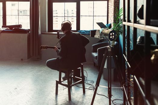 Close up of a man singer in a headphones with a guitar recording a track in a home studio. Man wearing sunglasses, jeans, black shirt and a jacket. Look from back, window on a bakground