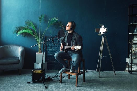 Close up of a man singer in a headphones with a guitar recording a track in a home studio. Man wearing sunglasses, jeans, black shirt and a jacket.