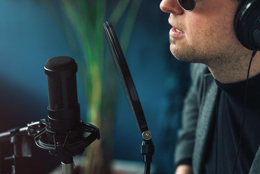 Close up of a man singer in a headphones recording a song in a home studio. Man wearing sunglasses, black shirt and a jacket. side view