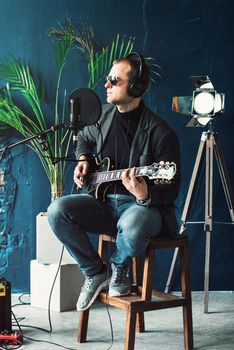 Close up of a man singer in a headphones with a guitar recording a track in a home studio. Man wearing sunglasses, jeans, black shirt and a jacket. side view