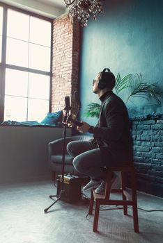 Close up of a man singer in a headphones with a guitar recording a track in a home studio. Man wearing sunglasses, jeans, black shirt and a jacket. Look from back, window on a bakground