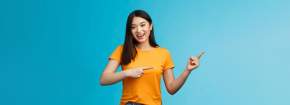 Upbeat friendly asian cute girl with dark haircut lively pointing left introduce awesome new copy space, smiling broadly toothy happy grin, stand blue background joyful, recommend link.