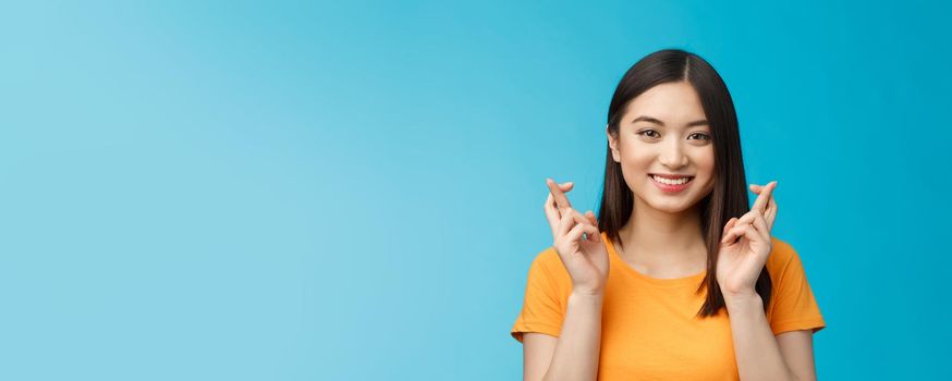 Close-up hopeful cute asian girl with short dark hair cross fingers good luck, praying smiling broadly awaiting positive news, faithfully hope win, stand blue background upbeat excited.