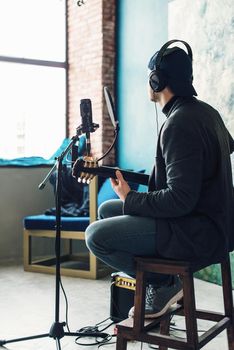 Close up of a man singer in a headphones with a guitar recording a track in a home studio. Man wearing jeans black shirt and a jacket. Look from back