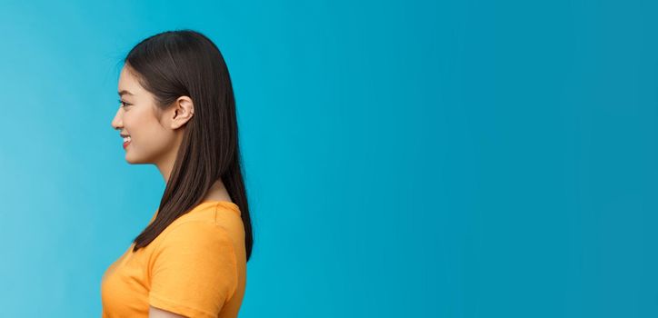 Profile shot asian cute woman perfect skin, turn left, smiling joyfully, pose near blue background, standing queue, awaiting coffee take-away, relaxed chatting girlfriend, wear yellow summer t-shirt.