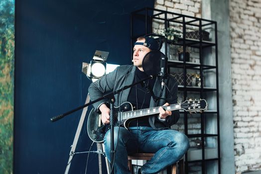 Close up of a man singer in a headphones with a guitar recording a track in a home studio. Man wearing jeans black shirt and a jacket.