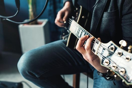 Close up of a man singer in a headphones with a guitar recording a track in a home studio. Man wearing sunglasses, jeans, black shirt and a jacket. side view