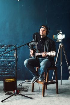 Close up of a man singer in a headphones with a guitar recording a track in a home studio. Man wearing jeans black shirt and a jacket.