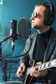 Close up of a man singer in a headphones with a guitar recording a track in a home studio. Man wearing sunglasses, jeans, black shirt and a jacket. side view