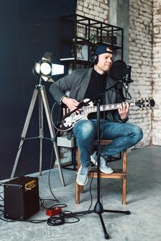 Close up of a man singer in a headphones with a guitar recording a track in a home studio. Man wearing jeans black shirt and a jacket.