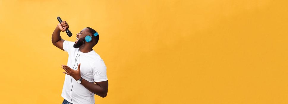 Portrait of cheerful positive chic. handsome african man holding microphone and having headphones on head listening music singing song enjoying weekend vacation isolated on yellow background.