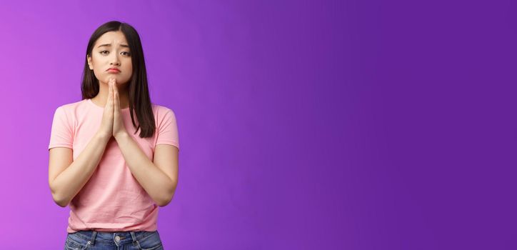 Gloomy cute asian daughter begging mother buy new phone, supplicating, hold hands pray, sulking frowning pretty please, asking apology, pleading favor, stand purple background. Copy space