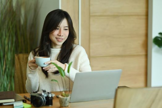 Smiling asian woman wearing warm sweater and drinking hot coffee while using laptop computer a home.