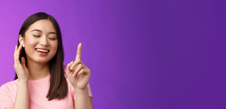 Close-up cheerful glad attractive asian woman close eyes raising one finger waiting chorus, smiling delighted touch wireless earphone, make volume louder satisfied enjoy songs, purple background.