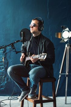 Close up of a man singer in a headphones with a guitar recording a track in a home studio. Man wearing sunglasses, jeans, black shirt and a jacket. side view