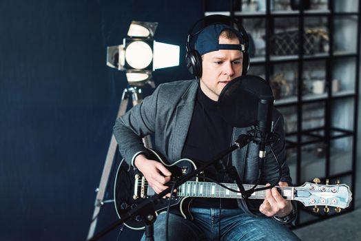 Close up of a man singer in a headphones with a guitar recording a track in a home studio. Man wearing jeans black shirt and a jacket.