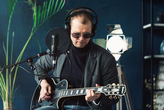 Close up of a man singer in a headphones with a guitar recording a track in a home studio. Man wearing sunglasses, jeans, black shirt and a jacket.