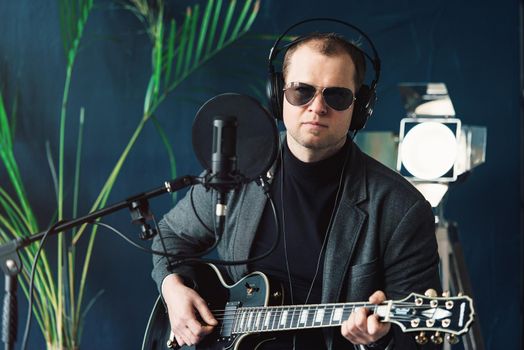 Close up of a man singer in a headphones with a guitar recording a track in a home studio. Man wearing sunglasses, jeans, black shirt and a jacket.