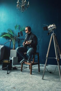 Close up of a man singer in a headphones with a guitar recording a track in a home studio. Man wearing sunglasses, jeans, black shirt and a jacket. side view