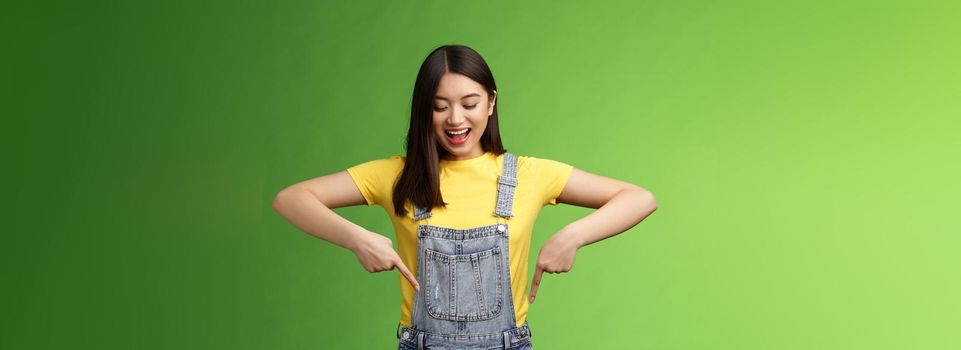 Impressed entertained cute asian girl watching pointing down interested, observe cool promo, smiling broadly, fascinated gaze bottom advertisement, take look curious thing, green background.