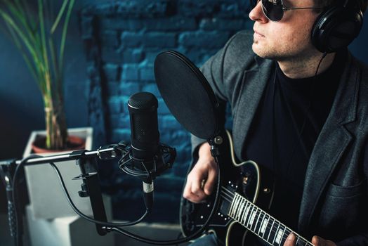 Close up of a man singer in a headphones with a guitar recording a track in a home studio. Man wearing sunglasses, jeans, black shirt and a jacket. side view