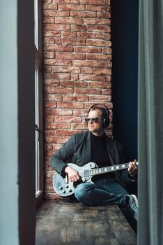 Man singer sitting on a window sill in a headphones with a guitar recording a track in a home studio. Man wearing sunglasses, jeans, black shirt and a jacket.