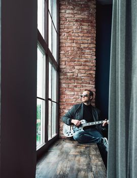 Man singer sitting on a window sill in a headphones with a guitar recording a track in a home studio. Man wearing sunglasses, jeans, black shirt and a jacket.