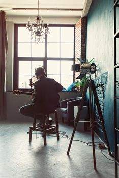 Close up of a man singer in a headphones with a guitar recording a track in a home studio. Man wearing sunglasses, jeans, black shirt and a jacket. Look from back, window on a bakground