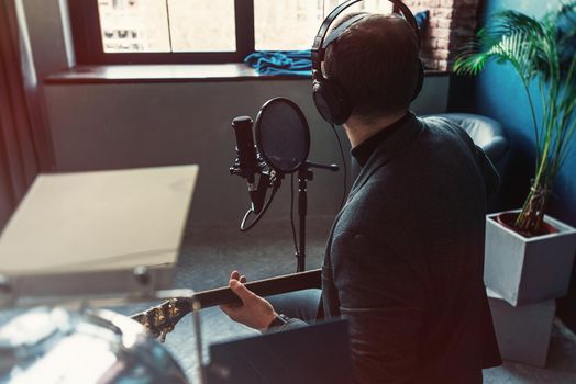 Close up of a man singer in a headphones with a guitar recording a track in a home studio. Man wearing sunglasses, jeans, black shirt and a jacket. Look from back, window on a bakground