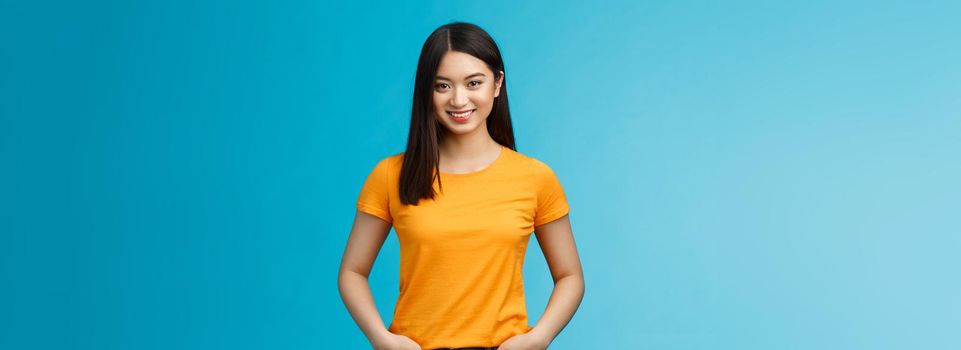 Confident assertive smiling asian woman stand blue background look camera, hold hands jeans pockets express self-assured vibe, enjoy seeing positive results skincare procedures, wear yellow t-shirt.